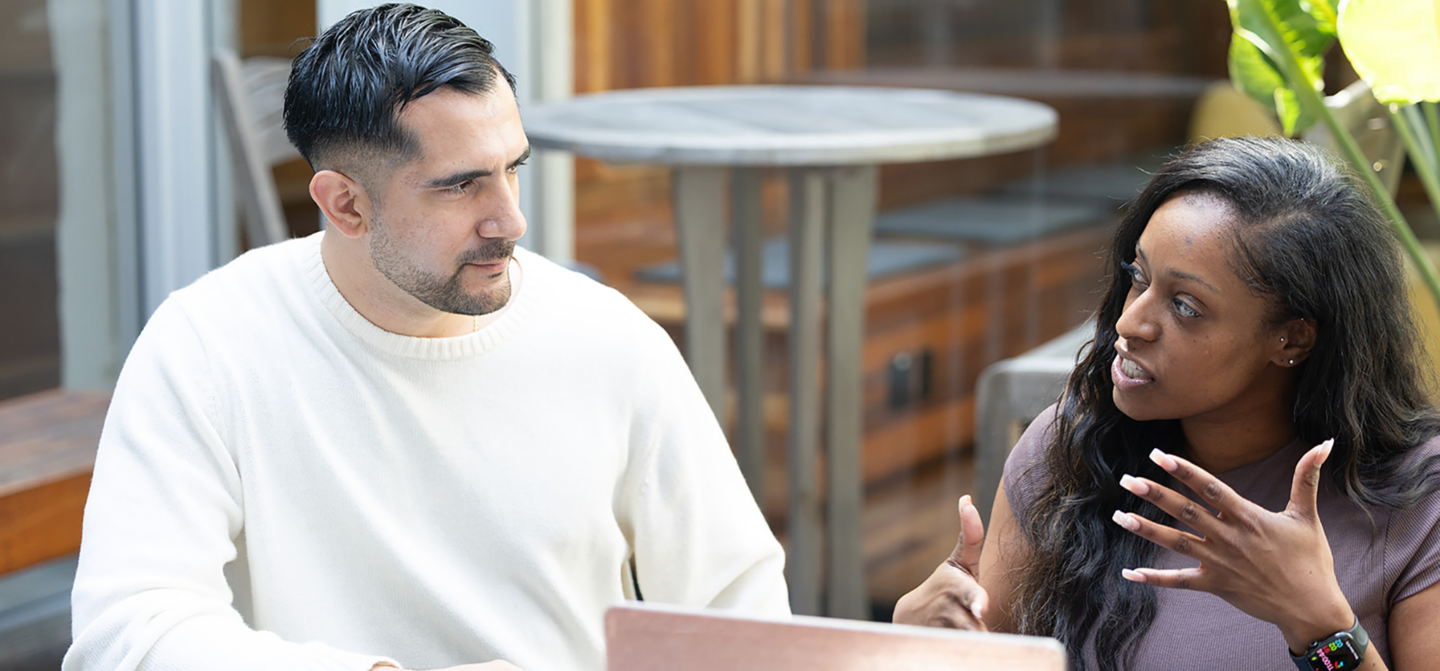 A woman and a man sit at a desk having a conversation in front of an open laptop.